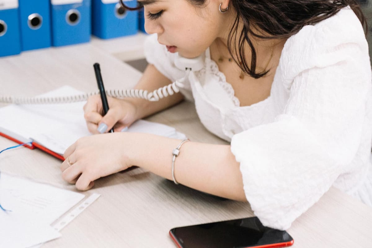 A woman takes notes while talking on the phone at a desk with document binders behind her.