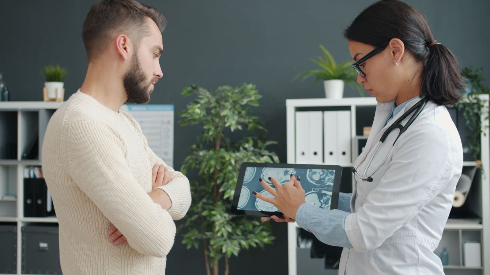 A physician in a white coat points to MRI images on a tablet while discussing them with a patient standing opposite her in a clinic office.