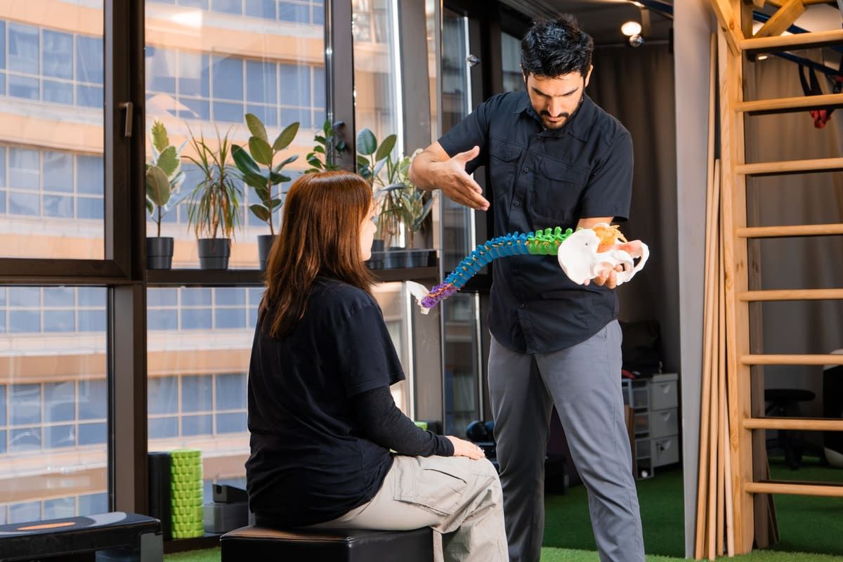 A clinician holds a colorful anatomical model of the spine and pelvis, gesturing as he explains it to a seated patient in a bright rehabilitation studio.
