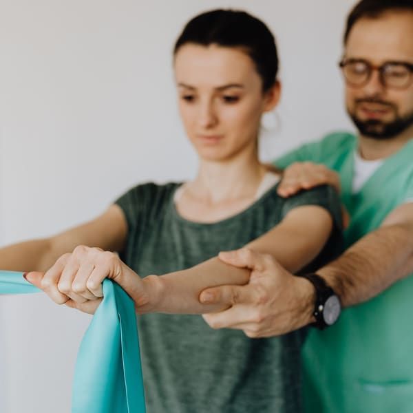 A clinician in green scrubs steadies a patient by the shoulder and forearm as she pulls a teal resistance band with both arms, an anatomy chart on the wall behind them.