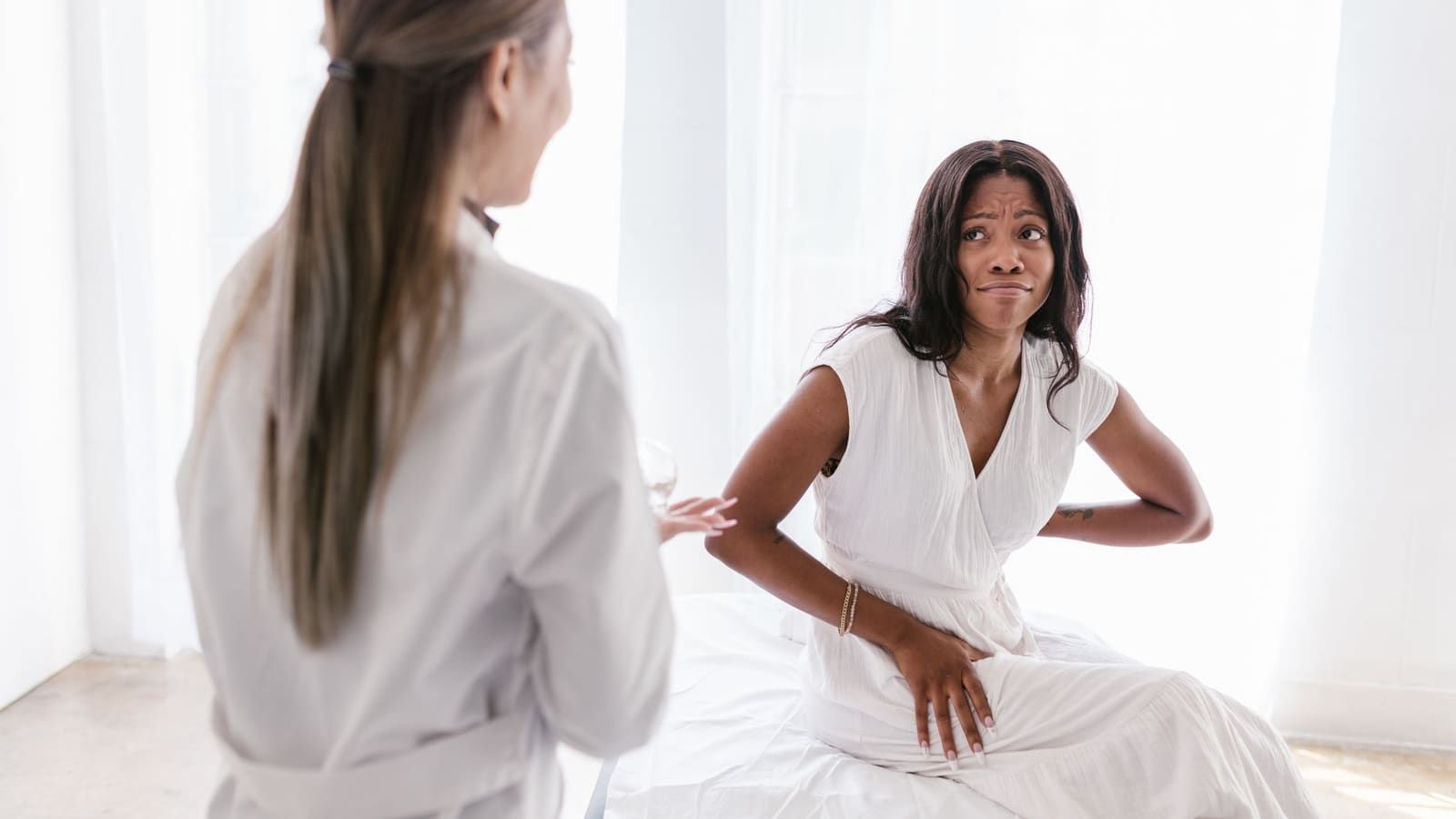 A patient sitting on an exam table rests one hand on her lower back and one on her thigh while describing her pain to a clinician in a white coat.
