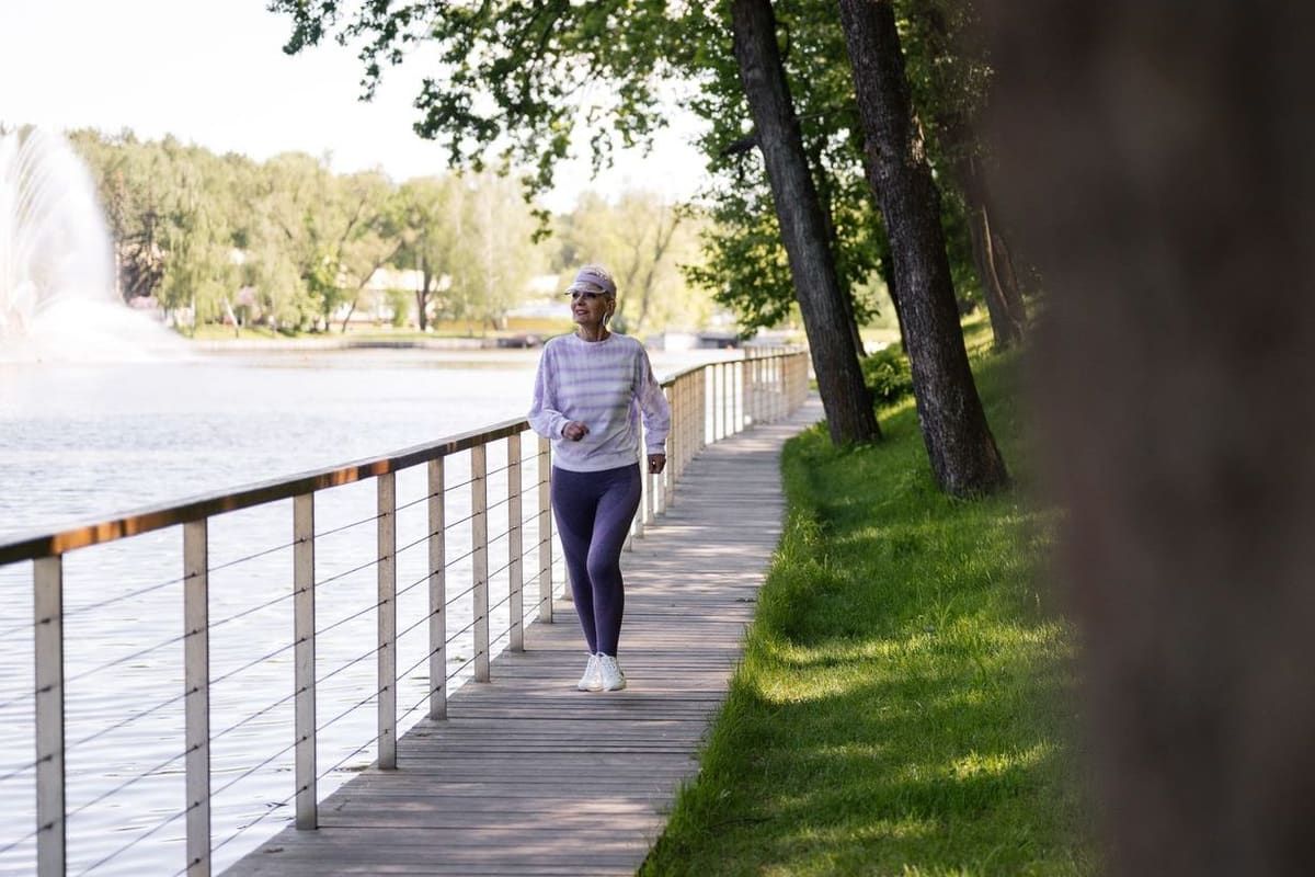 An older woman in walking clothes makes her way along a lakeside boardwalk on a sunny morning.