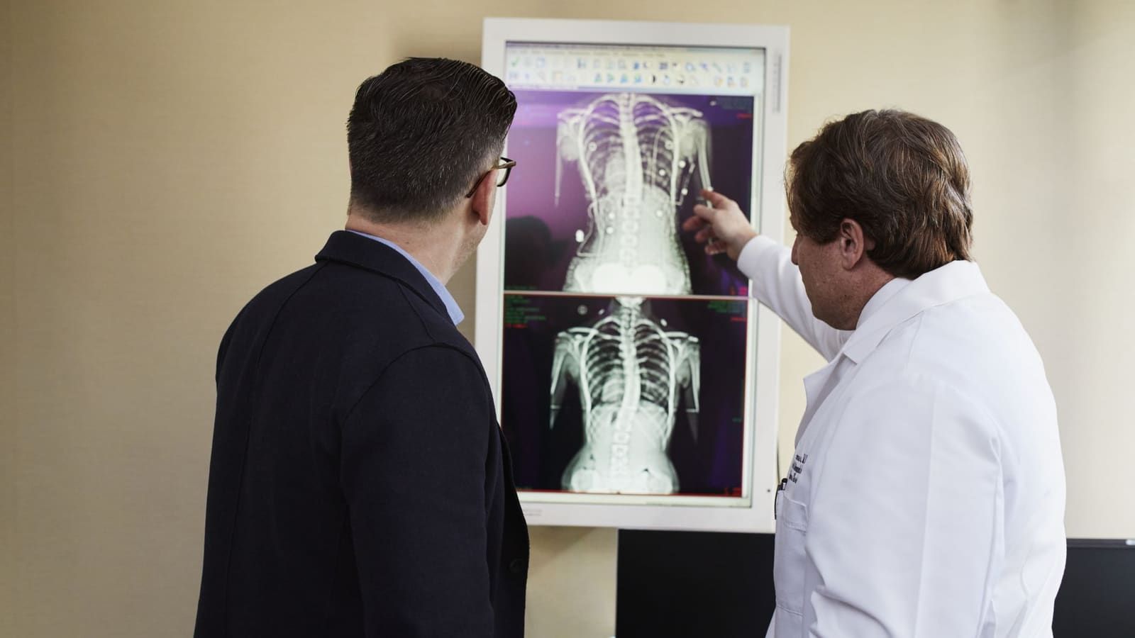 A physician in a white coat points to spine images on a wall-mounted monitor while a patient in a dark jacket stands beside him and studies the screen.