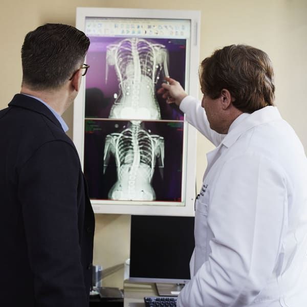 A physician in a white coat points to spine images on a wall-mounted monitor while a patient in a dark jacket stands beside him and studies the screen.